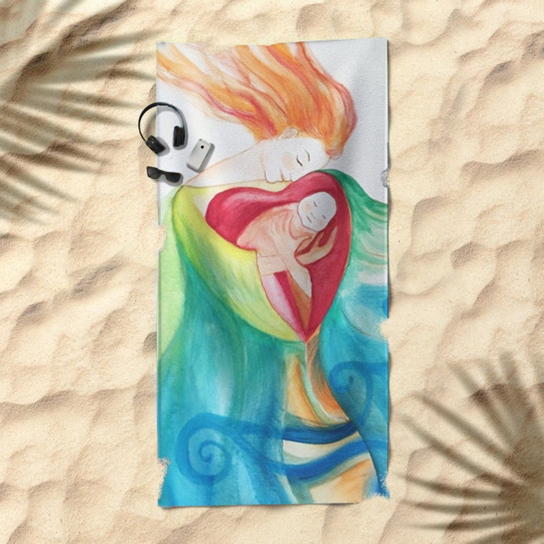May include: Beach towel featuring a watercolor illustration of a mother and child embraced within a heart shape. The artwork uses a palette of red, green, blue, and orange. The towel is rectangular and rests on a sandy surface.