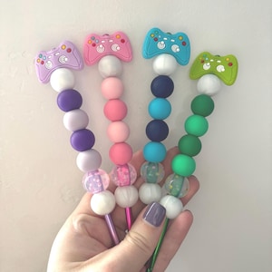 Peut inclure: Quatre crochets en métal avec des manches en perles de silicone. Chaque crochet est surmonté d'une breloque en forme de manette de jeu vidéo colorée. Les perles sont disposées dans des tons de violet, rose, bleu et vert, avec des finitions unies et pailletées.