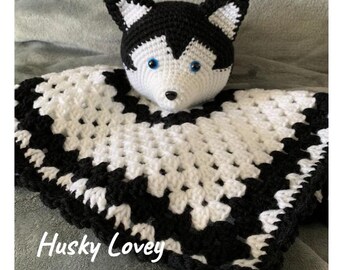 May include: A handmade crochet Husky lovey with a black and white color scheme. The Husky's head is white with black markings and blue eyes. The blanket portion is white with black edging. Text on the image reads "Husky Lovey Crochet Pattern."