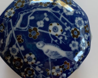 May include: A blue ceramic trinket box with a floral and bird design. The lid features a white bird and white and brown flowers against a blue background. The box has a unique, rounded shape.