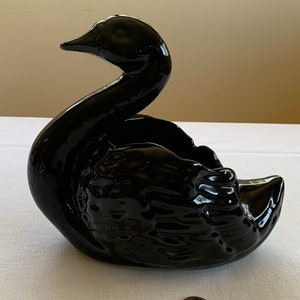 Puede incluir: Un macetero de cerámica negro en forma de cisne con un acabado brillante. El cuello del cisne está curvado y su cabeza está inclinada hacia un lado. El macetero mide aproximadamente 18 cm de largo.