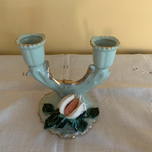 May include: A light blue ceramic candle holder with two candle cups and a white rose with green leaves. The holder has a gold trim.