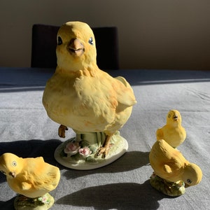 Könnte beinhalten: Ein Set aus drei gelben Keramik-Kükenfiguren. Das größte Küken steht aufrecht mit ausgebreiteten Flügeln. Die beiden kleineren Küken sitzen. Alle drei Küken haben schwarze Augen und Schnäbel.