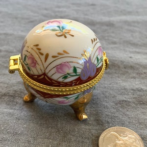 Puede incluir: Una pequeña caja de porcelana blanca en forma de huevo con una tapa con bisagras de oro. La caja está decorada con flores rosas y moradas y detalles dorados. La caja se apoya en tres patas doradas.