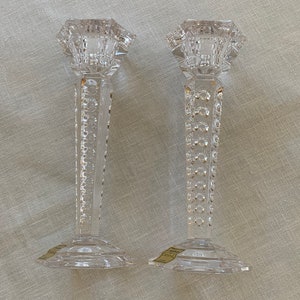K&ouml;nnte beinhalten: Zwei klare Glaskerzenhalter mit geometrischem Design. Die Kerzenhalter haben einen quadratischen Sockel und einen hohen, schlanken Stiel mit einem dekorativen Muster. Die Oberseite jedes Kerzenhalters ist ein Quadrat mit einer kleinen &Ouml;ffnung f&uuml;r eine Kerze.
