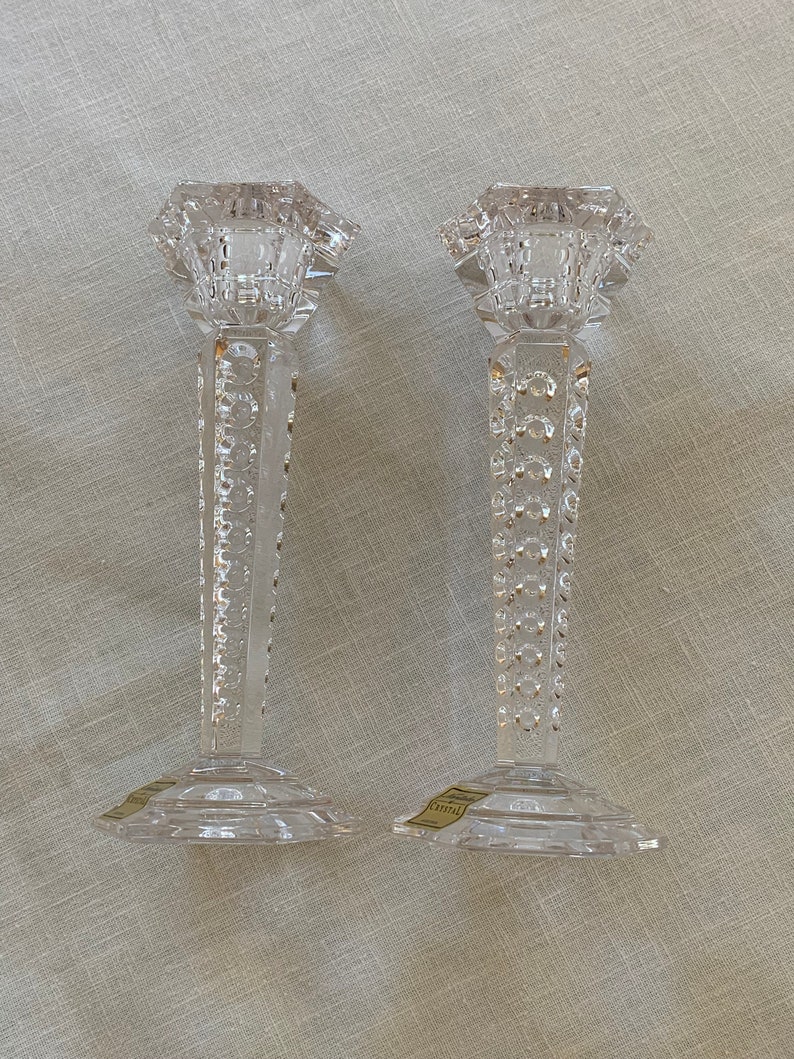 K&ouml;nnte beinhalten: Zwei klare Glaskerzenhalter mit geometrischem Design. Die Kerzenhalter haben einen quadratischen Sockel und einen hohen, schlanken Stiel mit einem dekorativen Muster. Die Oberseite jedes Kerzenhalters ist ein Quadrat mit einer kleinen &Ouml;ffnung f&uuml;r eine Kerze.