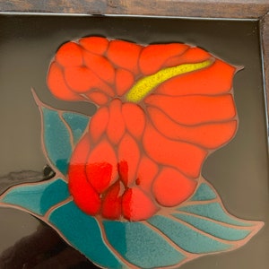 Può includere: Un fiore rosso e giallo con foglie verdi su uno sfondo nero. Il fiore è in una cornice di piastrelle in ceramica.