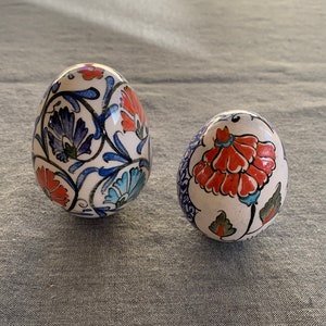 Può includere: Due ornamenti in ceramica a forma di uovo. Ogni uovo è bianco con un motivo floreale rosso, blu e verde. Le uova sono su una superficie grigia. I motivi floreali includono fiori e foglie stilizzate.
