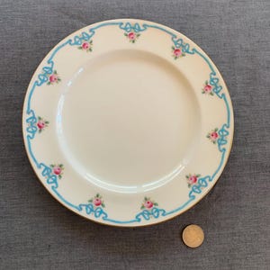 Könnte beinhalten: Ein weißer Keramik-Dinnerteller mit blauem und goldenem Rand. Der Teller hat ein florales Design mit rosa Rosen und blauen Bändern.