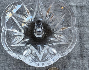 May include: A clear glass ring holder with a starburst pattern. The holder has a raised center post with a small ring.