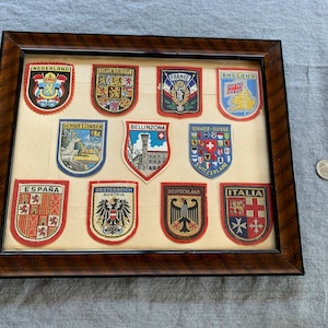 May include: A framed collection of 12 vintage travel patches featuring national symbols and crests from countries including Netherlands, Belgium, France, England, Switzerland, Spain, Austria, Germany, and Italy.