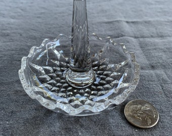 Op de afbeelding: Een glazen ringhouder van helder glas met een verhoogde middelste paal en een gegolfde rand. De houder staat op een grijs stoffen oppervlak.