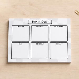 May include: A white notepad with the words "Brain Dump" at the top. The notepad is divided into six sections labeled "Must Do," "Could Do," "Want to Do," "Call," "Schedule," and "Message." A gold paperclip rests on the wooden surface.