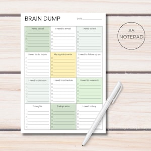 Pode incluir: Bloco de notas A5 com as palavras "BRAIN DUMP" no topo. O bloco de notas tem seções para anotações, incluindo "I need to call", "I need to email" e "I need to text". Uma caneta branca repousa no bloco de notas.