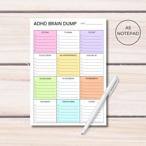 Puede incluir: Bloc de notas A5 con el texto "ADHD BRAIN DUMP" y secciones para tareas como "To Call", "To Email" y "To Buy". El bloc de notas tiene un diseño colorido con un bolígrafo blanco encima.