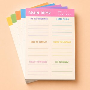 May include: A stack of rectangular paper notepads with a pink header labeled BRAIN DUMP and sections for My Top Priorities, I Need To Do, I Need To Contact, I Need To Schedule, I Need To Purchase, and For Tomorrow, with colorful edges in yellow, green, orange, and blue.