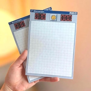 May include: Two rectangular paper notepads featuring a Minesweeper game interface design. The top header includes a blue title bar, a yellow smiley face icon, and red digital score displays. The main body consists of a white grid on a gray background.