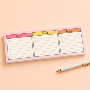 May include: A rectangular notepad with a pink border featuring three columns labeled Do Now, Do next, and Do Later, each with lined spaces and checkboxes. A white ballpoint pen with gold-colored accents is positioned next to the notepad.