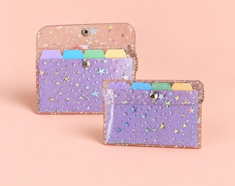 May include: Two rectangular clear plastic pouches with a glittery star pattern and a snap button closure. Each pouch contains four colorful index tabs in purple, blue, green, and yellow, with a purple insert visible inside the clear material.