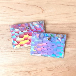 May include: Two rectangular pouches made of a clear, iridescent material featuring a geometric, mosaic-like pattern of polygons. Each pouch has a flap closure secured with a single silver-toned metal snap button, reflecting pink, blue, and yellow hues.