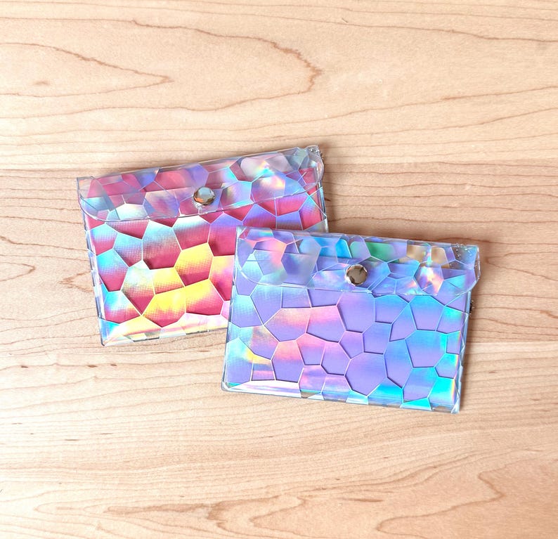 May include: Two rectangular pouches made of a clear, iridescent material featuring a geometric, mosaic-like pattern of polygons. Each pouch has a flap closure secured with a single silver-toned metal snap button, reflecting pink, blue, and yellow hues.