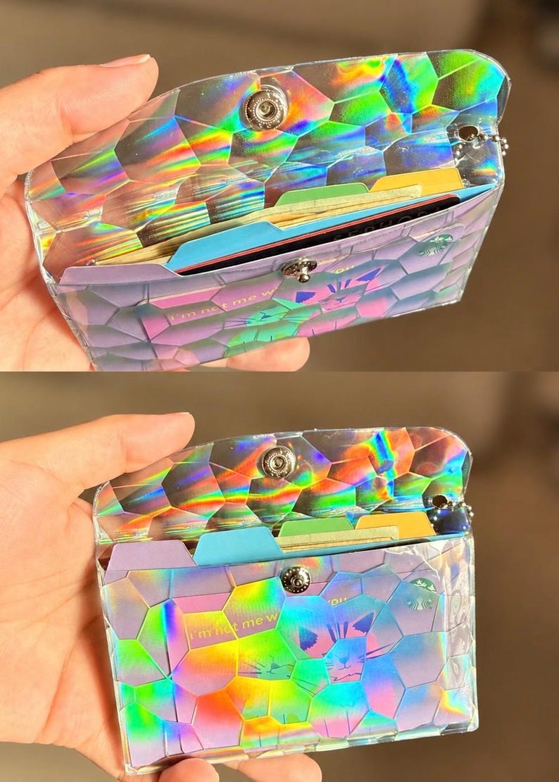 May include: A holographic wallet with a geometric hexagonal pattern and a metallic snap closure. The wallet contains multiple colorful dividers and cards, including one with the visible text 'I'm not me w' and a small circular green logo.