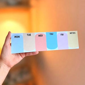 May include: A rectangular weekly planner notepad featuring six vertical sections with rounded bottoms in light blue, beige, pink, bright blue, lavender, and yellow. Each section is labeled with black text: MON, TUE, WED, THU, FRI, and NOTES.