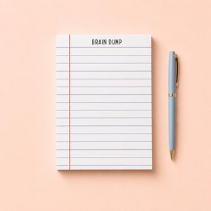 May include: A white notepad with horizontal blue lines, a vertical red margin line, and the text BRAIN DUMP printed at the top, accompanied by a light blue ballpoint pen with a gold clip and tip, positioned to the right of the notepad.
