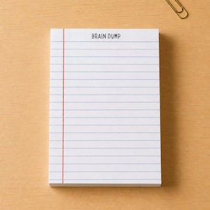 May include: A white notepad with blue horizontal lines and a red vertical line on the left side. The words "BRAIN DUMP" are printed at the top. A gold paperclip rests on a light brown wooden surface.