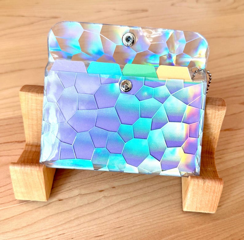 May include: A small, rectangular, iridescent wallet with a geometric, stained-glass-like pattern in shades of purple, blue, and green. The wallet features a clear, holographic outer layer, a silver metal snap closure, and internal dividers.