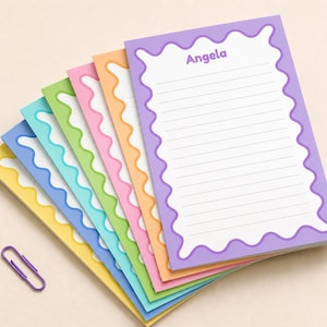 May include: A stack of colorful notepads with wavy borders in yellow, blue, green, pink, orange, and purple. The top notepad is purple with the name "Angela" printed at the top. A purple paperclip rests near the bottom.