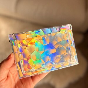 May include: A rectangular, clear pouch featuring a geometric, shattered-glass pattern that reflects iridescent, holographic colors including yellow, orange, blue, and green. The pouch has a stitched border and a small metal zipper pull on the side.