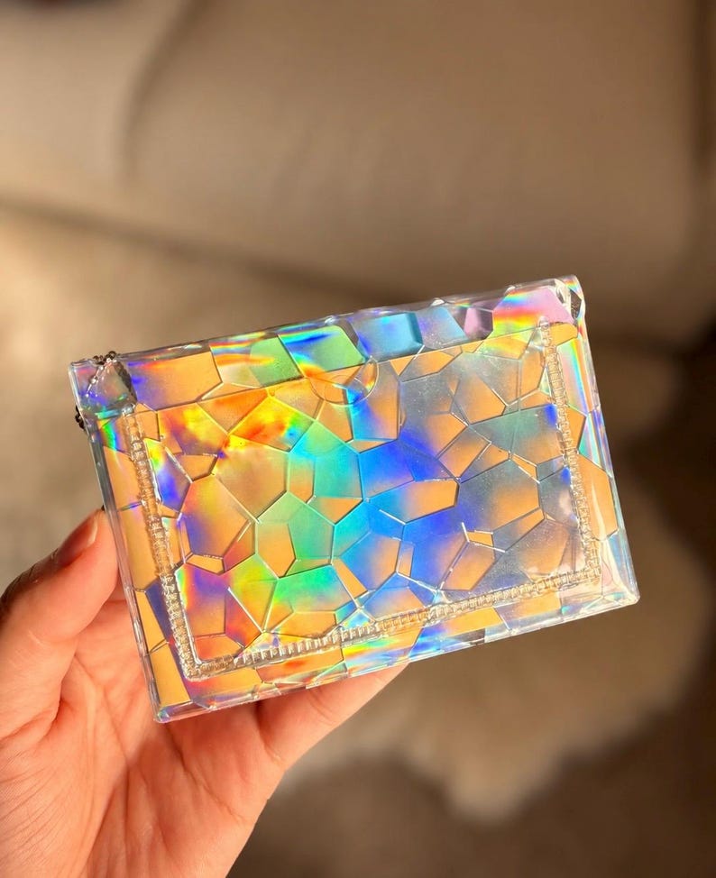 May include: A rectangular, clear pouch featuring a geometric, shattered-glass pattern that reflects iridescent, holographic colors including yellow, orange, blue, and green. The pouch has a stitched border and a small metal zipper pull on the side.