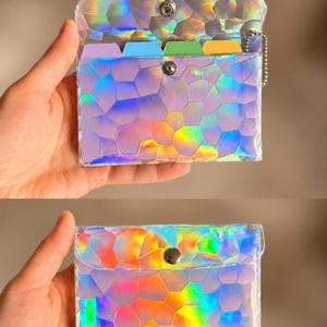 May include: A rectangular holographic card holder with a geometric mosaic pattern that reflects rainbow colors. The accessory features a metal snap button closure, a silver ball chain attachment, and internal dividers in purple, blue, green, and yellow.