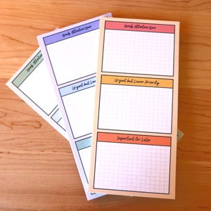 May include: Several rectangular notepads with grid lines and color-coded sections. The notepads have sections labeled "Needs Attention Now", "Urgent but Lower Priority", and "Important for Later". The notepads are on a wooden surface.