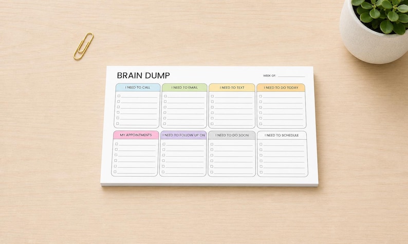 K&ouml;nnte beinhalten: Ein wei&szlig;er Brain Dump Notizblock mit Abschnitten f&uuml;r Aufgaben wie Anrufe, E-Mails und Termine. Der Notizblock hat ein farbenfrohes Layout mit Text wie "I need to call" und "I need to do today". Eine goldene B&uuml;roklammer liegt auf der Holzoberfl&auml;che.