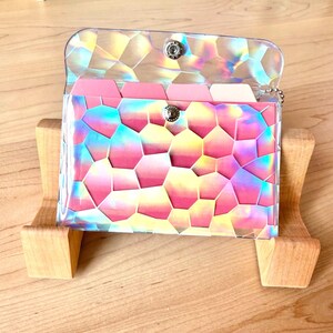 May include: A rectangular card holder featuring a clear, iridescent, geometric mosaic pattern overlay. The interior contains multiple pink and white tabbed dividers. The case is secured with a silver-toned metal snap button closure at the center.