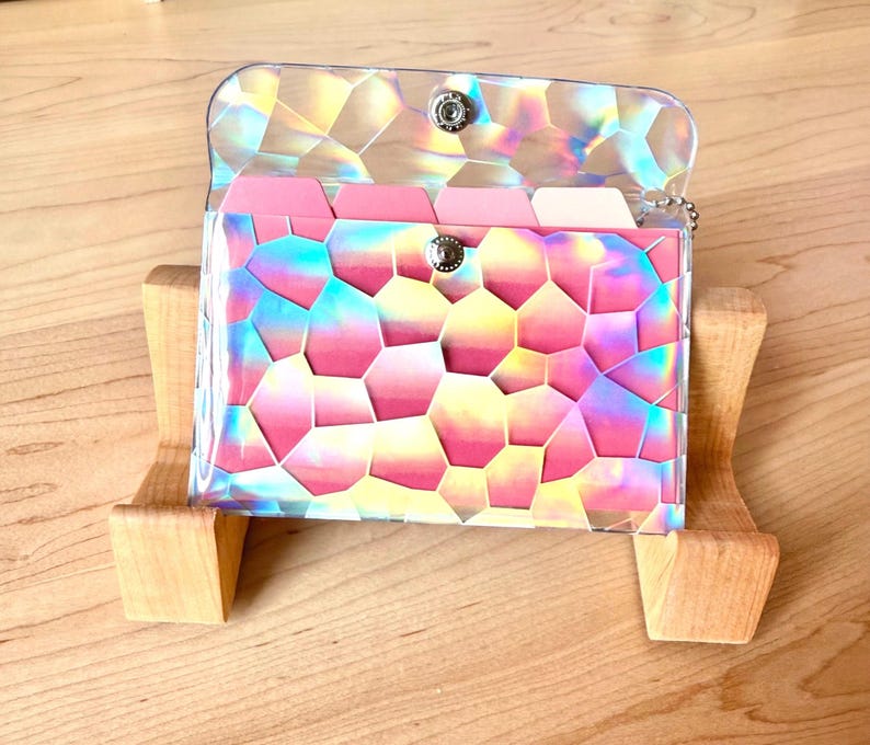 May include: A rectangular card holder featuring a clear, iridescent, geometric mosaic pattern overlay. The interior contains multiple pink and white tabbed dividers. The case is secured with a silver-toned metal snap button closure at the center.