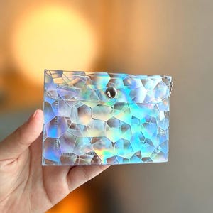 May include: A rectangular pouch featuring a translucent, iridescent material with a geometric, multi-faceted pattern that reflects various colors. The pouch has a flap closure secured by a single silver-toned metal snap button at the center.