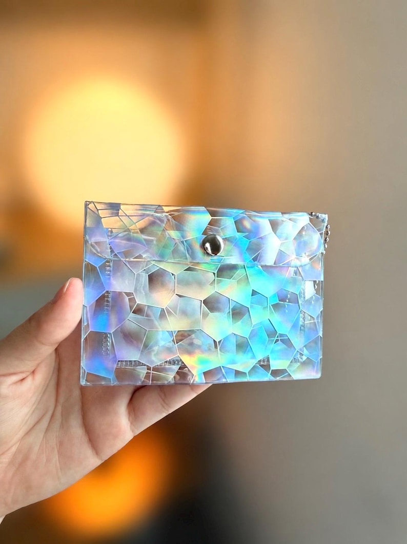 May include: A rectangular pouch featuring a translucent, iridescent material with a geometric, multi-faceted pattern that reflects various colors. The pouch has a flap closure secured by a single silver-toned metal snap button at the center.