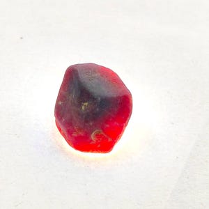May include: A small, raw, unpolished gemstone with a vibrant red hue. The stone features an irregular, rounded shape with a darker, opaque patch near the top center and translucent, bright red edges that catch the light, showing natural texture.