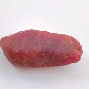 Puede incluir: Una gema de rubí en bruto sin cortar. El rubí es de color rojo intenso con una forma ligeramente irregular y una superficie texturizada. La gema se muestra sobre un fondo blanco.