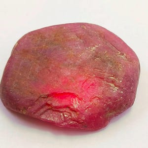 Puede incluir: Una gema de rubí en bruto, sin cortar, de color rojo intenso. La superficie tiene un aspecto texturizado con sutiles variaciones de color, que van del rojo oscuro a tonos rosados más claros. La gema tiene forma irregular.