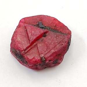 May include: A deep red ruby gemstone with a rough, natural surface. The ruby has a triangular shape and is a vibrant, rich red color.