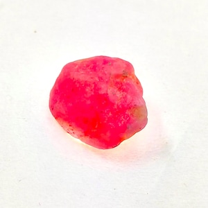 May include: A small, raw, unpolished gemstone with an irregular, rounded shape. The stone features a vibrant, saturated hot pink color throughout its surface, with subtle variations in tone and texture visible across its opaque, natural form.