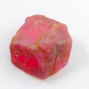 Puede incluir: Una piedra preciosa en bruto y sin pulir con un tono rosa y rojizo vibrante. La piedra presenta una forma irregular y maciza con facetas naturales visibles, superficies texturizadas y una profunda saturación de color en su estructura.