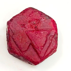 May include: A deep red ruby gemstone with a hexagonal shape and a faceted surface.