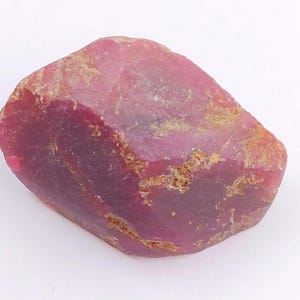 May include: A raw ruby gemstone, displaying a rich red colour. The surface has a textured appearance with lighter pink and brown patches. The irregularly shaped gemstone appears to be in its natural state.