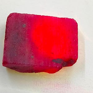 Puede incluir: Una gema rectangular en bruto, de color rojo vibrante. La piedra tiene un aspecto pulido y brillante en un lado, que pasa a una superficie más oscura y texturizada en el otro. Es probable que la gema sea un rubí.