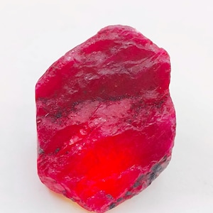 May include: A raw ruby gemstone in a deep red colour. The natural stone has a textured surface and an irregular shape, with areas of lighter and darker red. The gemstone is set against a white background.